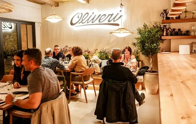 restaurant-empuriabrava-oliveri-ambiente-1 Interior acogedor del restaurant en Empuriabrava Oliveri, mostrando comensales disfrutando en mesas bajo iluminación cálida de lámparas de ratán y con un gran cartel luminoso de 'Oliveri' en la pared de fondo.